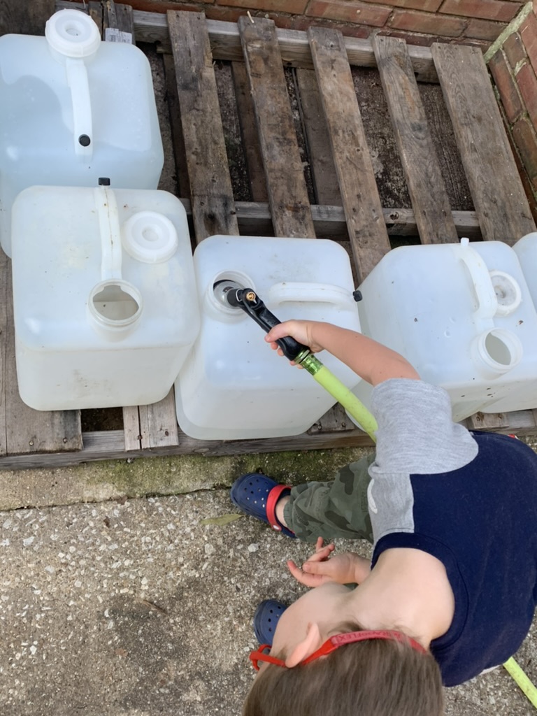 Child filling a large jug with a garden hose checking water level.